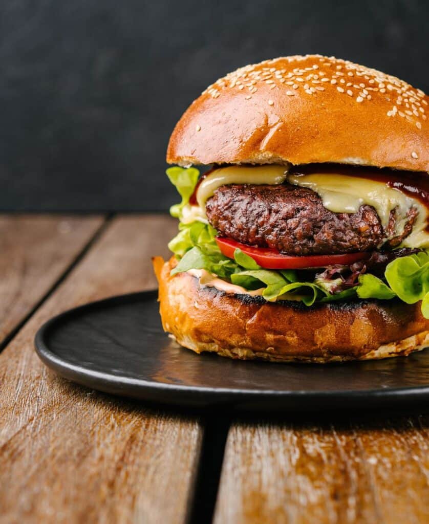 A mouthwatering vegan burger served on a black plate in La Plagne. The burger features a golden sesame seed bun, a plant-based patty with a slightly charred exterior, melted vegan cheese, fresh lettuce, tomato slices, and a drizzle of sauce. Set against a rustic wooden table, the gourmet burger looks rich, juicy, and indulgent.