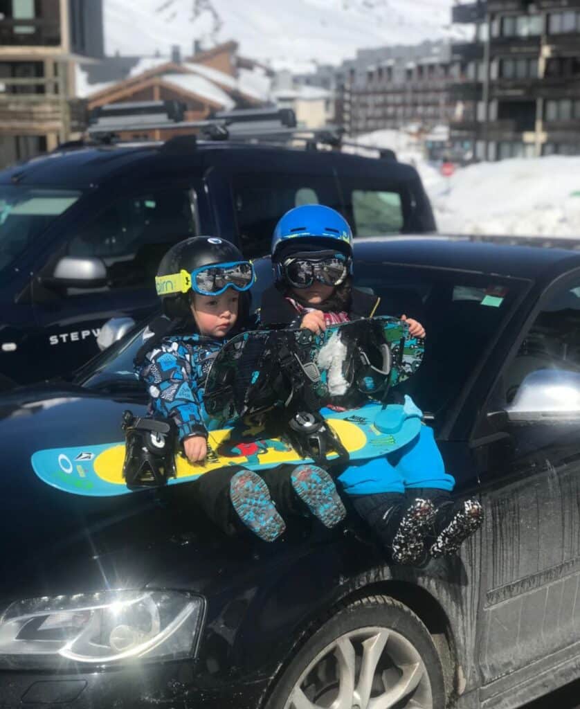 Two young children in colorful ski gear sit on the hood of a black car, each holding a snowboard. They wear helmets, ski goggles, and snow boots, with a snowy alpine village and parked cars in the background. Their expressions show excitement as they prepare for a day on the slopes.