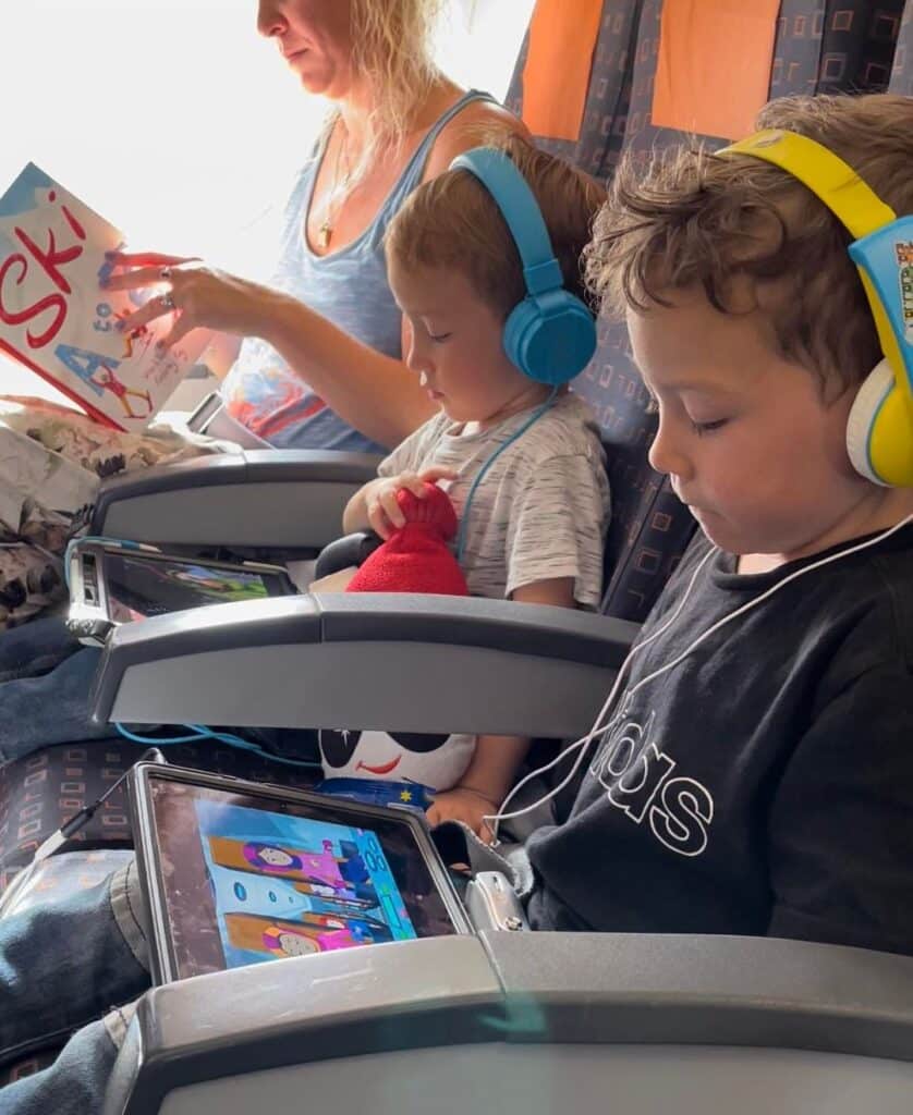 Two young boys sit side by side on an airplane, both wearing headphones and watching animated shows on tablets. One child clutches a plush toy with a red hat, while a woman seated next to them reads a ski-themed book. A cozy travel moment on the way to a winter destination.