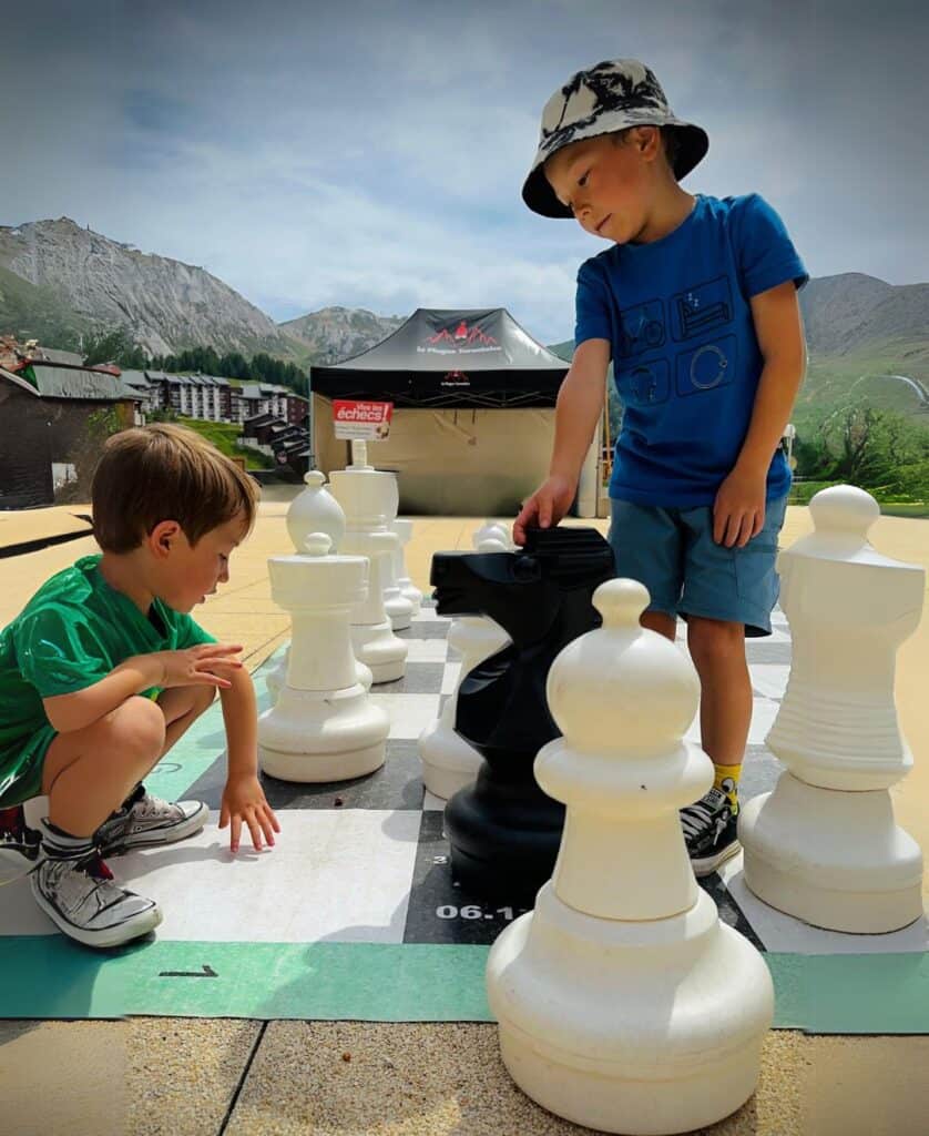Two young boys play an oversized outdoor chess game in La Plagne during a summer activity session, with scenic mountains in the background.