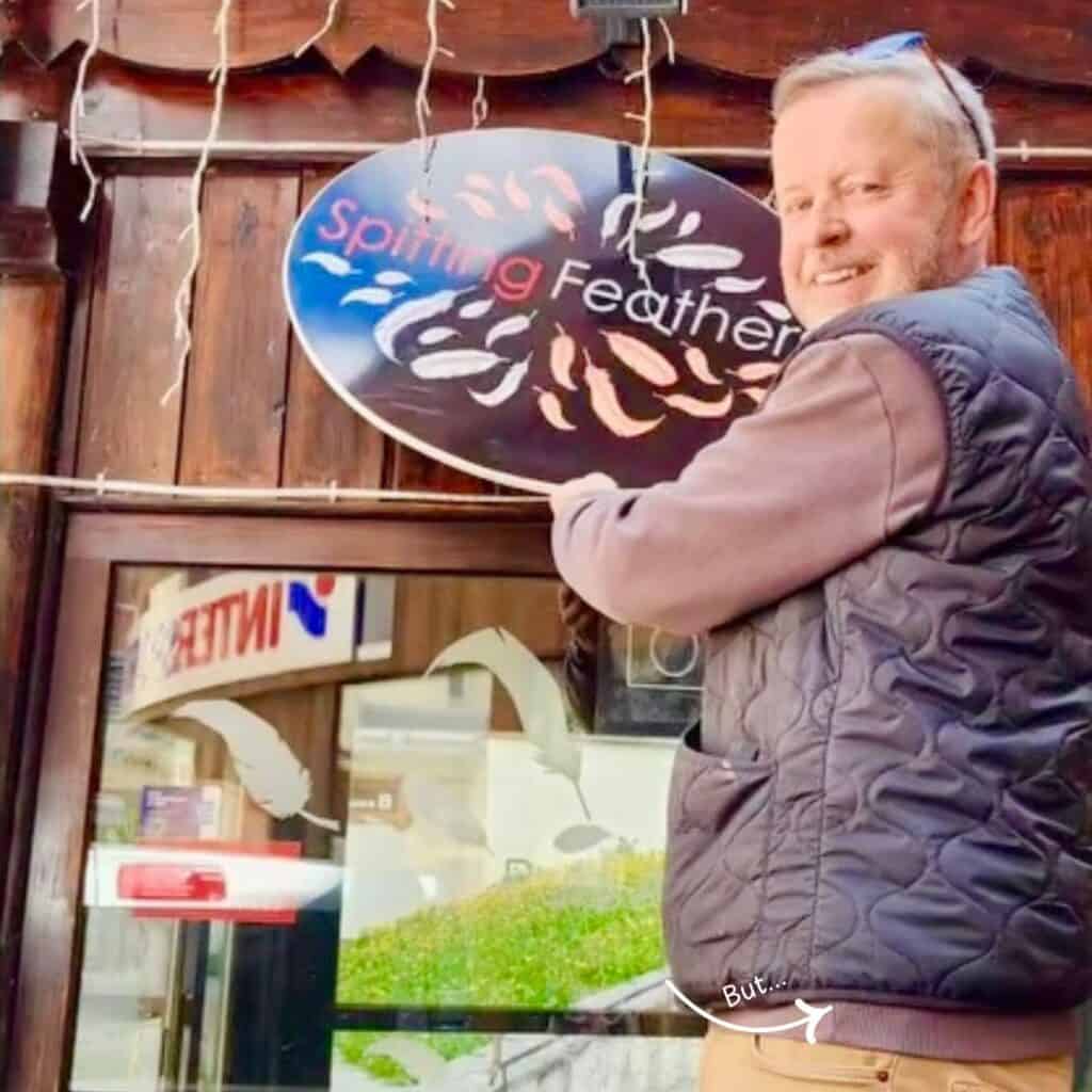 A man (Tim) smiling at the camera while taking down the old “Spitting Feathers” sign from the front of a wooden building, signaling the bar’s transition to new ownership. Reflections in the window show feather decals and a mirrored INTERSPORT sign, adding context to the original decor.