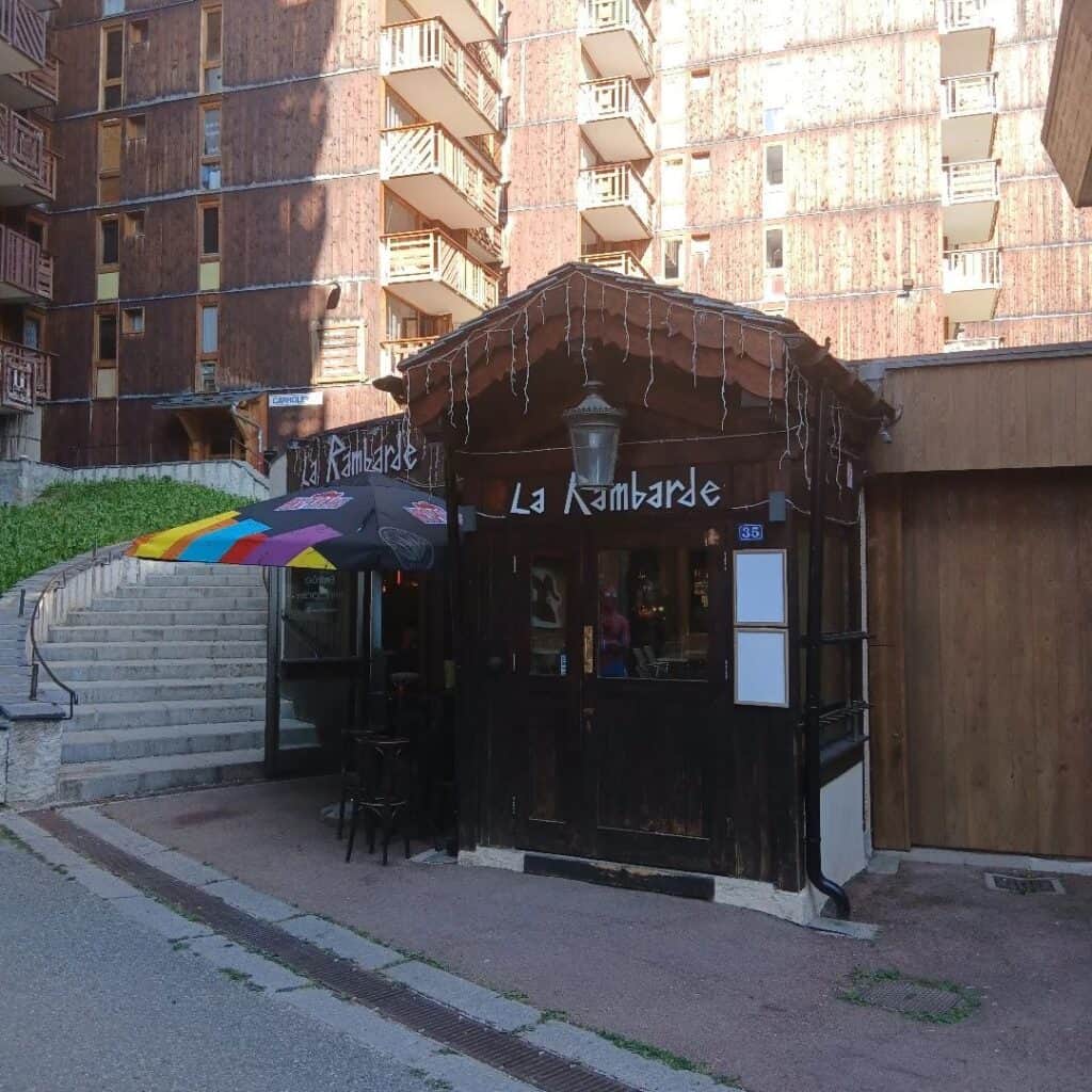 Street view of the newly named “La Rambarde” bar, with rustic wooden siding, a colorful outdoor umbrella, and white string lights hanging from the awning. The new signage is clearly visible above the entrance.