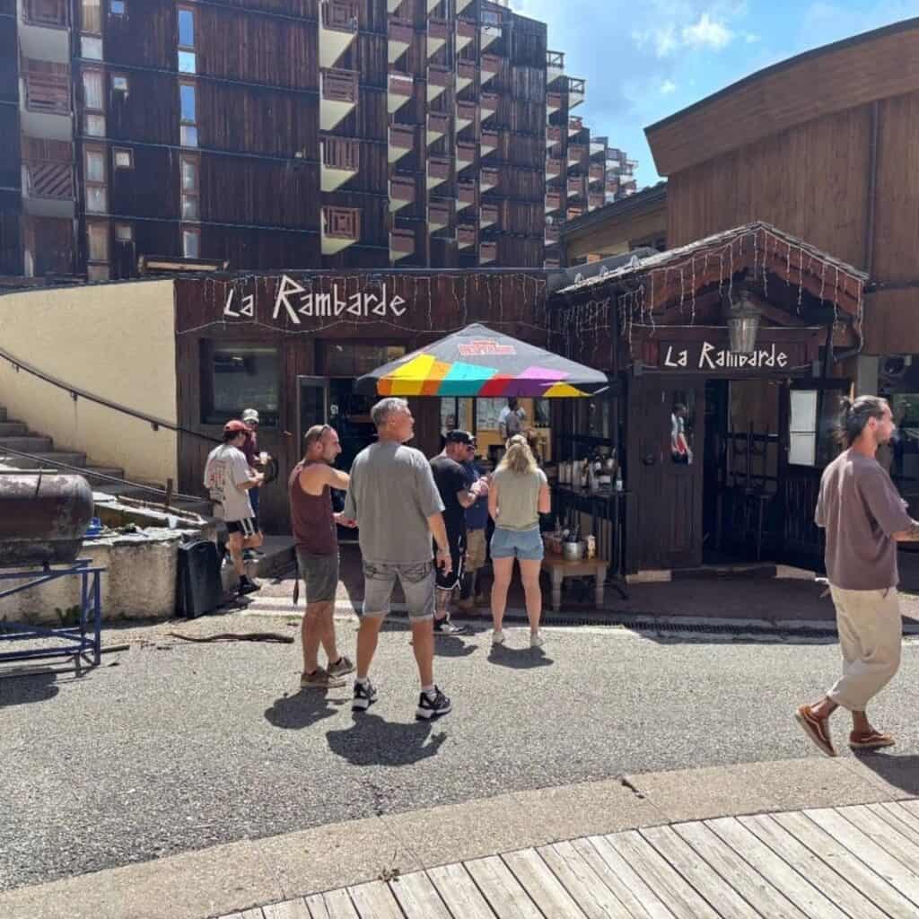 Street view of the newly named “La Rambarde” bar, with rustic wooden siding, a colorful outdoor umbrella, and white string lights hanging from the awning. The new signage is clearly visible above the entrance.