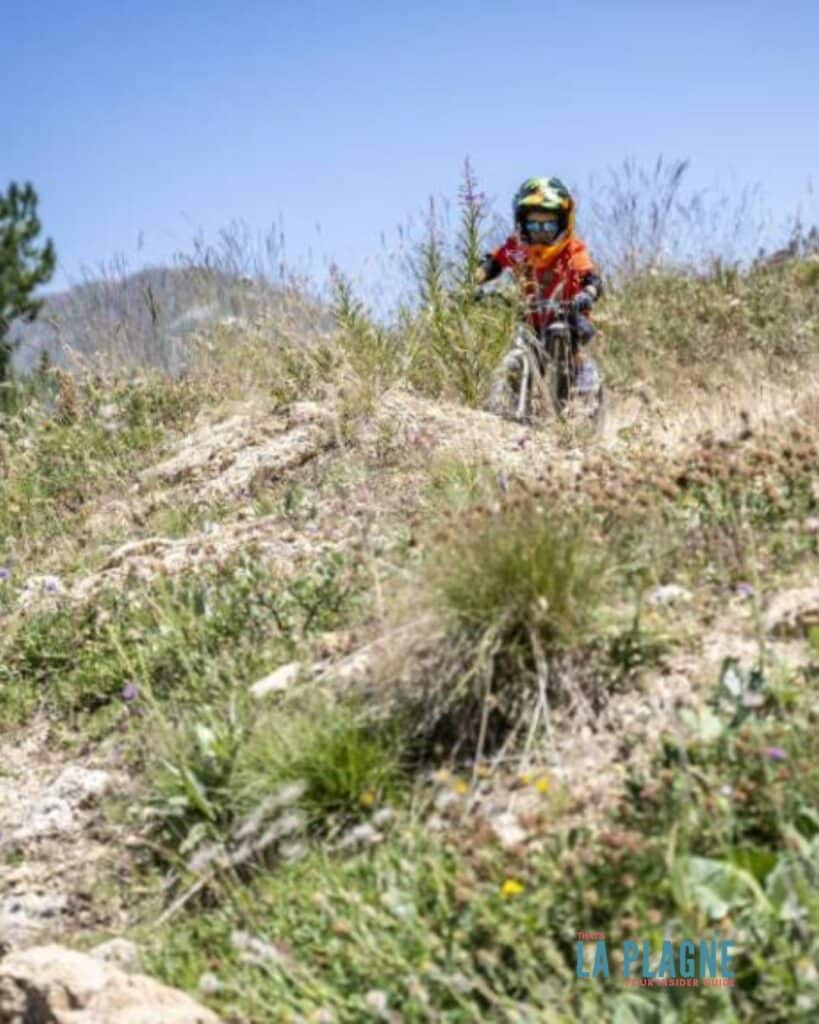 A small child (Odin from That's La Plagne) on a mountain bike rides through a rough, rocky trail surrounded by dry grass and wild plants, emphasizing the rugged natural terrain of La Plagne’s biking areas.
