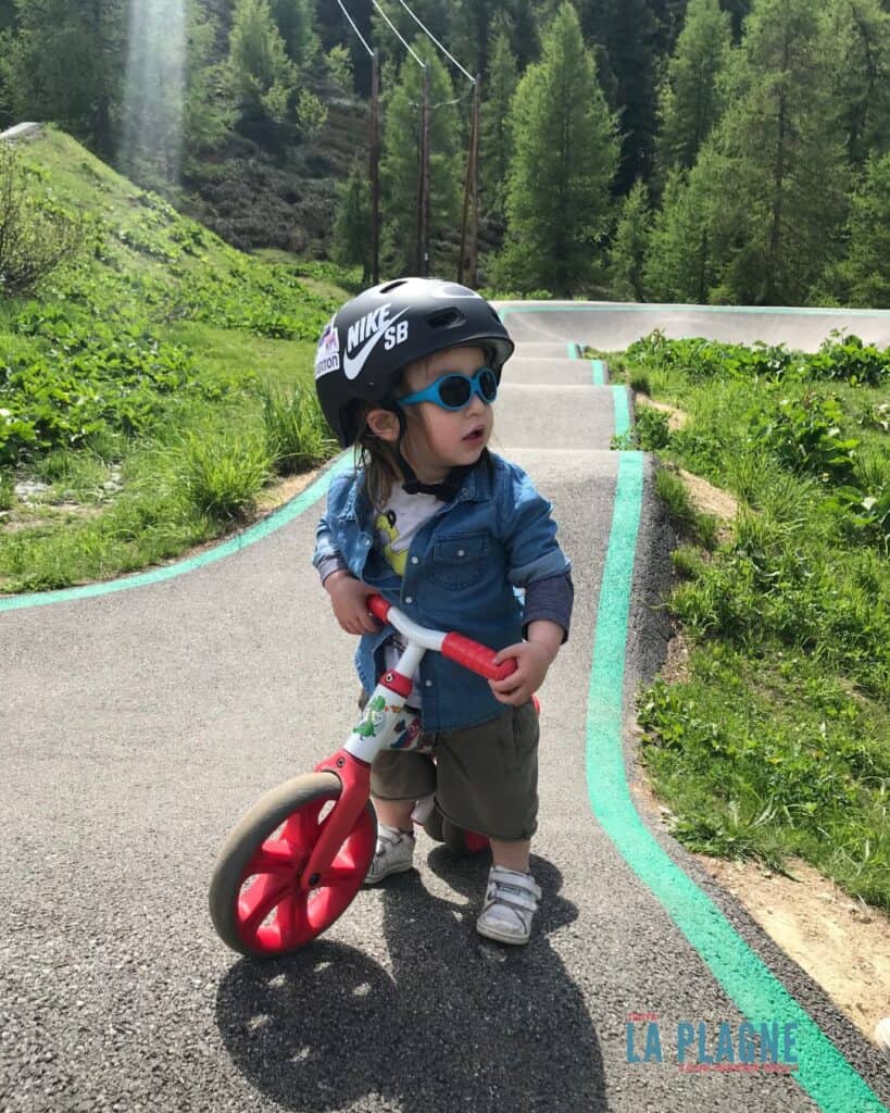 A toddler in sunglasses and a helmet stands beside a red balance bike on a paved pump track in Bellecote surrounded by lush greenery, highlighting La Plagne’s biking fun for even the youngest riders.