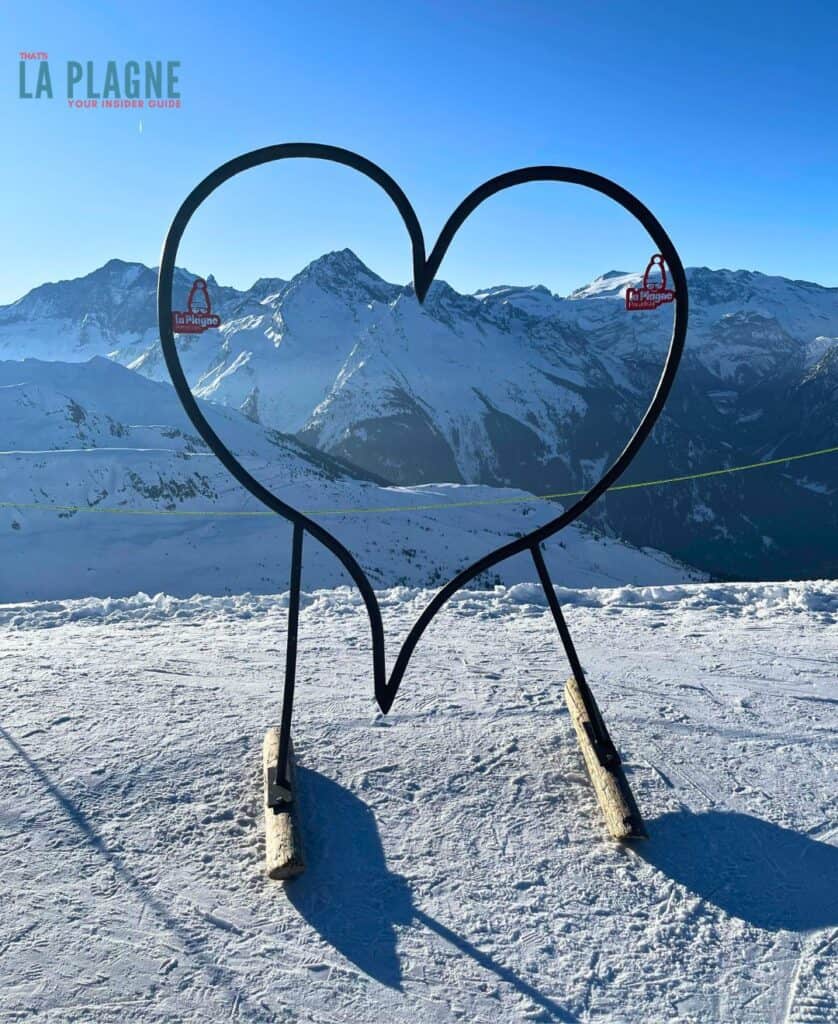 A large metal heart frame stands at a scenic overlook in La Plagne, perfectly framing snow-capped mountains — a charming photo op at the top of the grande rochette, that showcases the resort’s appeal for beginner skiers seeking a picturesque experience.