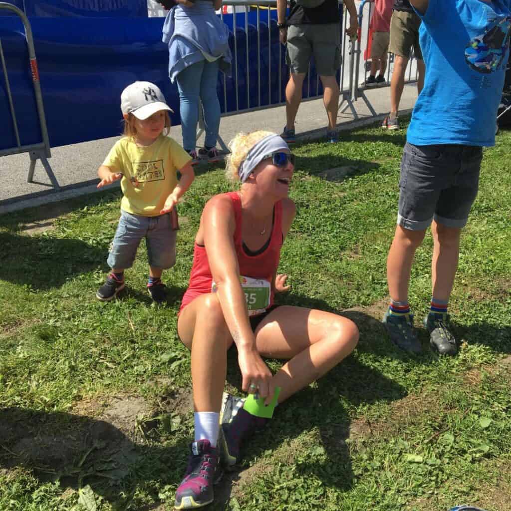 A sweaty, smiling woman in race gear sits on the grass after finishing her run, joined by a small child standing nearby. The joyful moment captures the family-friendly atmosphere of the 6000D La Plagne event.