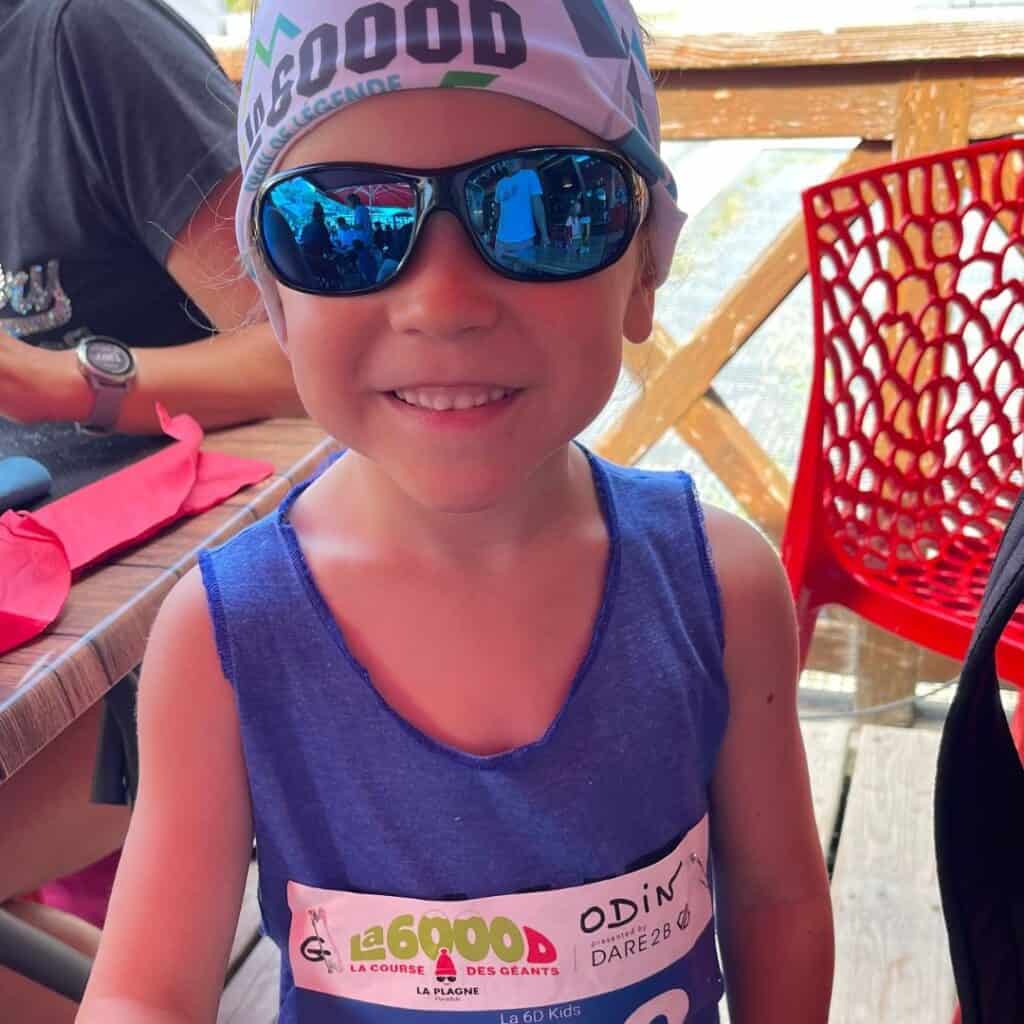 Close-up of a smiling child wearing a 6000D La Plagne bandana and sunglasses, proudly displaying a race bib that reads “La 6D Kids.” The image shows the inclusive and fun vibe of the youth race category.