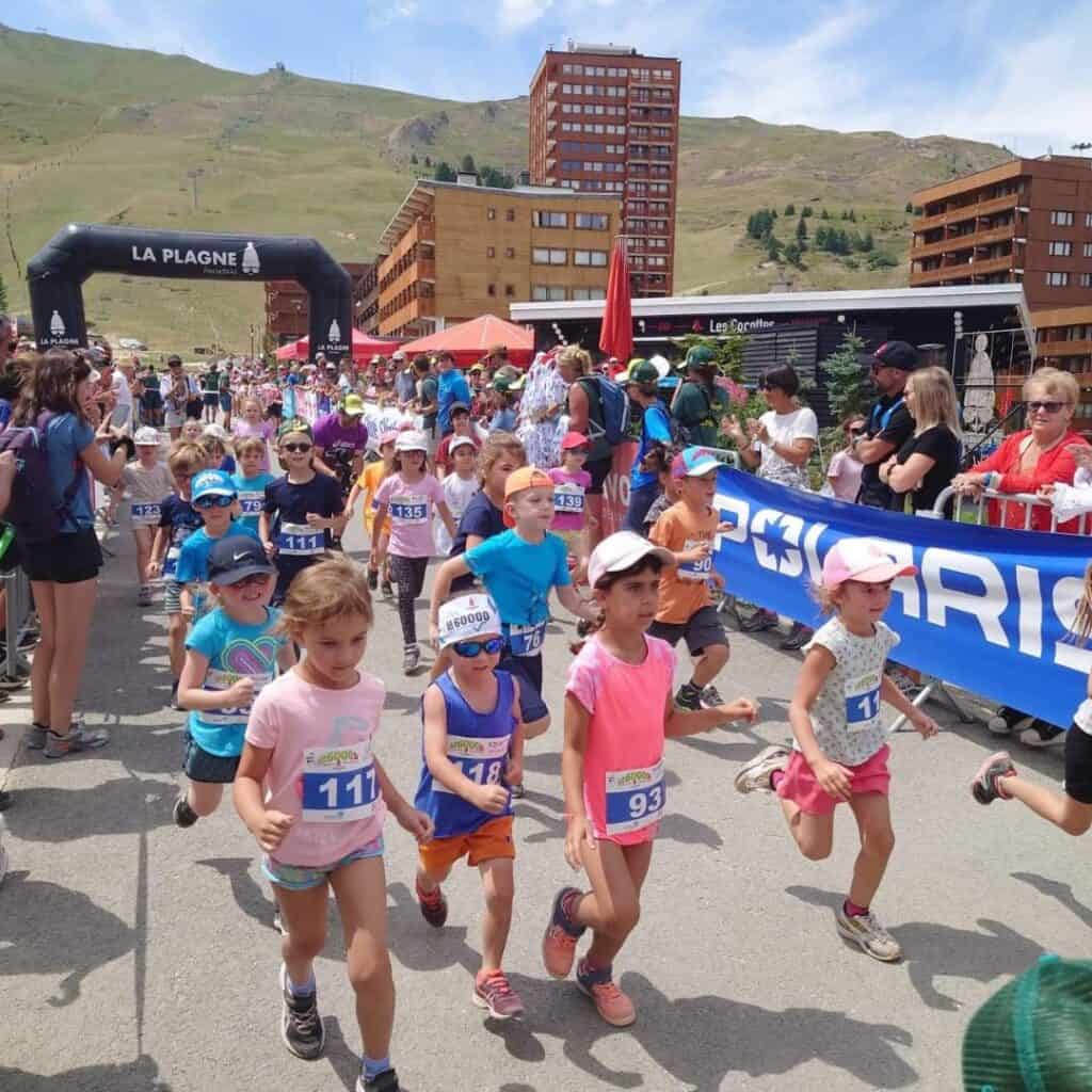 A large group of children take off at the start of the 6D Kids race during the 6000D La Plagne event, surrounded by cheering spectators and alpine buildings. The vibrant scene showcases the excitement and community energy of the race day.