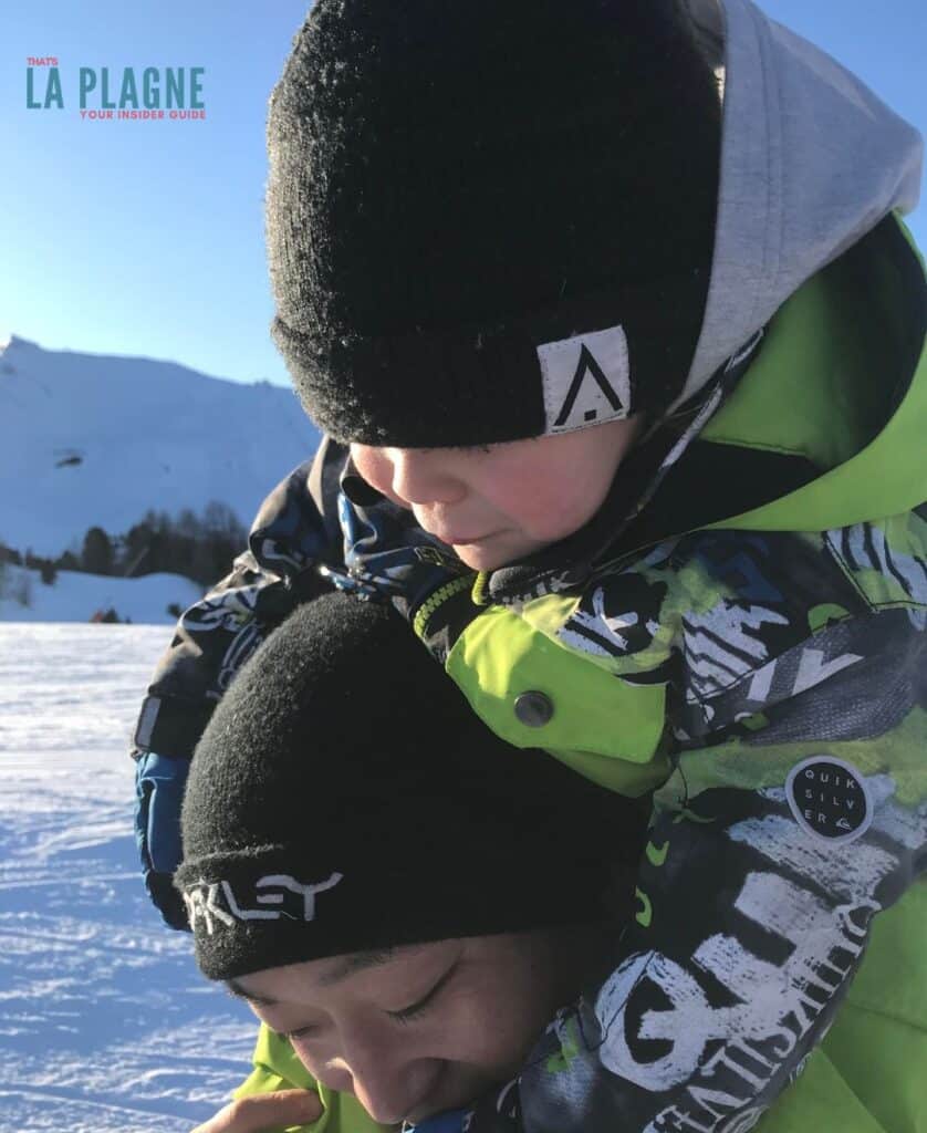 A father carrying his son in ski gear play and laugh together in the snow at La Plagne, capturing the family-friendly atmosphere perfect for first-timers and young learners.