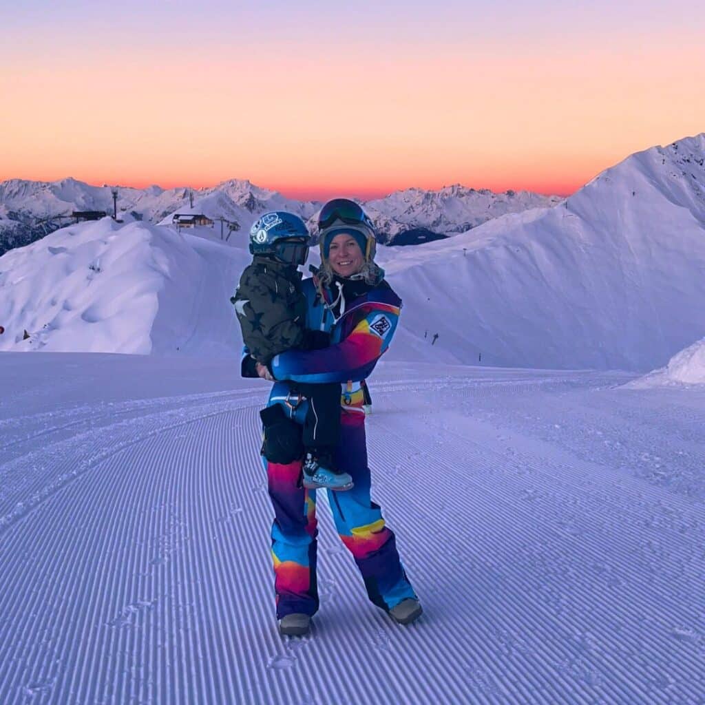 A person wearing a vibrant, rainbow-colored ski suit holds a child dressed in winter gear on a freshly groomed ski slope in La Plagne. The snowy mountains stretch behind them under a stunning sunrise, casting warm hues across the sky. Taken during the First Tracks activity, this moment captures the beauty of early morning skiing in the French Alps.