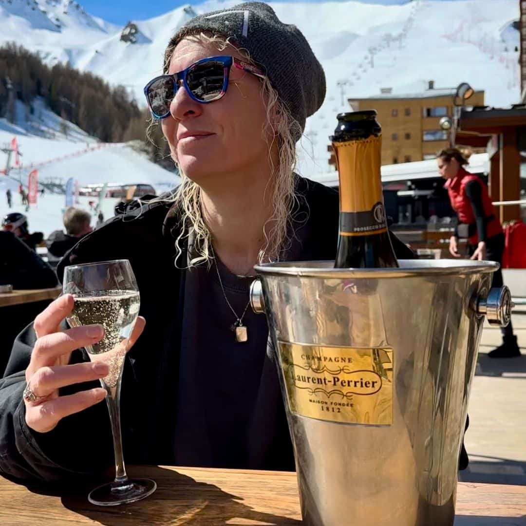 A woman in sunglasses and a beanie relaxes at an outdoor ski resort patio, holding a glass of champagne. A bottle of Laurent-Perrier champagne chills in an ice bucket on the wooden table beside her, with snow-covered mountains and ski slopes in the background.