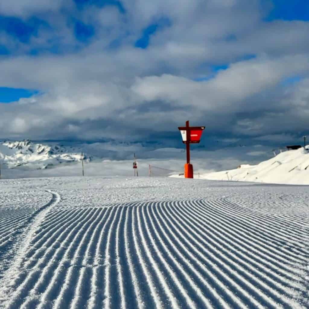 A freshly groomed ski slope with perfectly carved corduroy lines stretching into the distance under a blue sky with scattered clouds. A red and white signpost stands on the right, with snow-covered mountain peaks in the background.