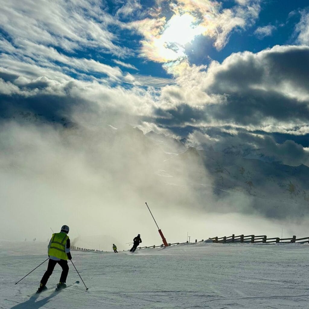 Skiers gliding down a misty slope in La Plagne as the sun breaks through thick clouds, casting a dramatic glow over the snow-covered peaks. One skier in a high-visibility vest leads the way down the freshly groomed run.