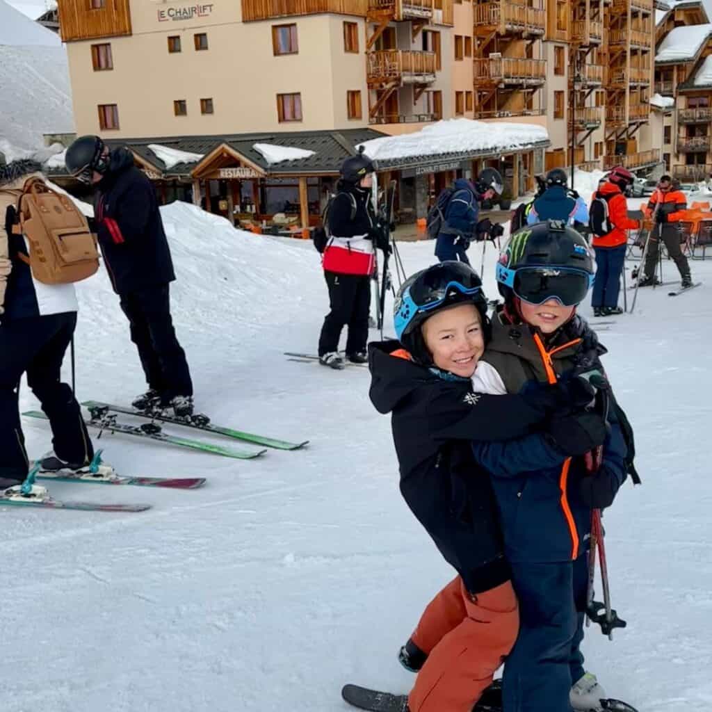 Two children in ski gear hugging joyfully on a snowy street in La Plagne, with a bustling ski village and wooden chalet-style buildings in the background. Other skiers are seen preparing their gear and socializing near a restaurant.