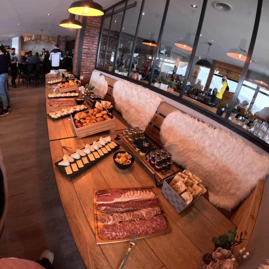 A beautifully arranged breakfast buffet inside a cozy mountain restaurant, featuring a spread of cheeses, charcuterie, croissants, jams, and fresh bread on a rustic wooden table with fluffy white cushions lining the bench. Warm lighting adds to the inviting atmosphere.