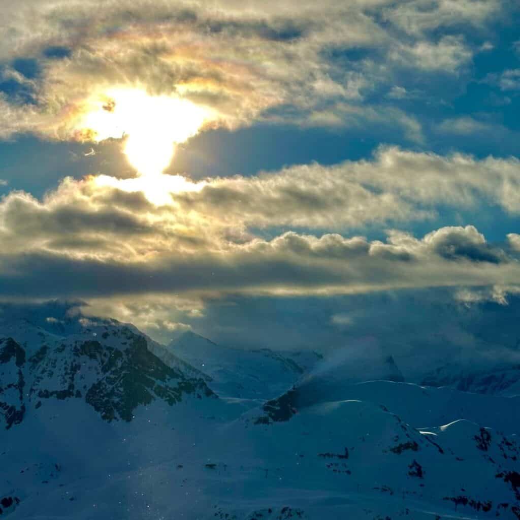 The sun breaking through dramatic clouds, casting a golden glow over the snowy mountain peaks in La Plagne. A faint rainbow-like halo forms around the sun, creating a breathtaking winter scene.