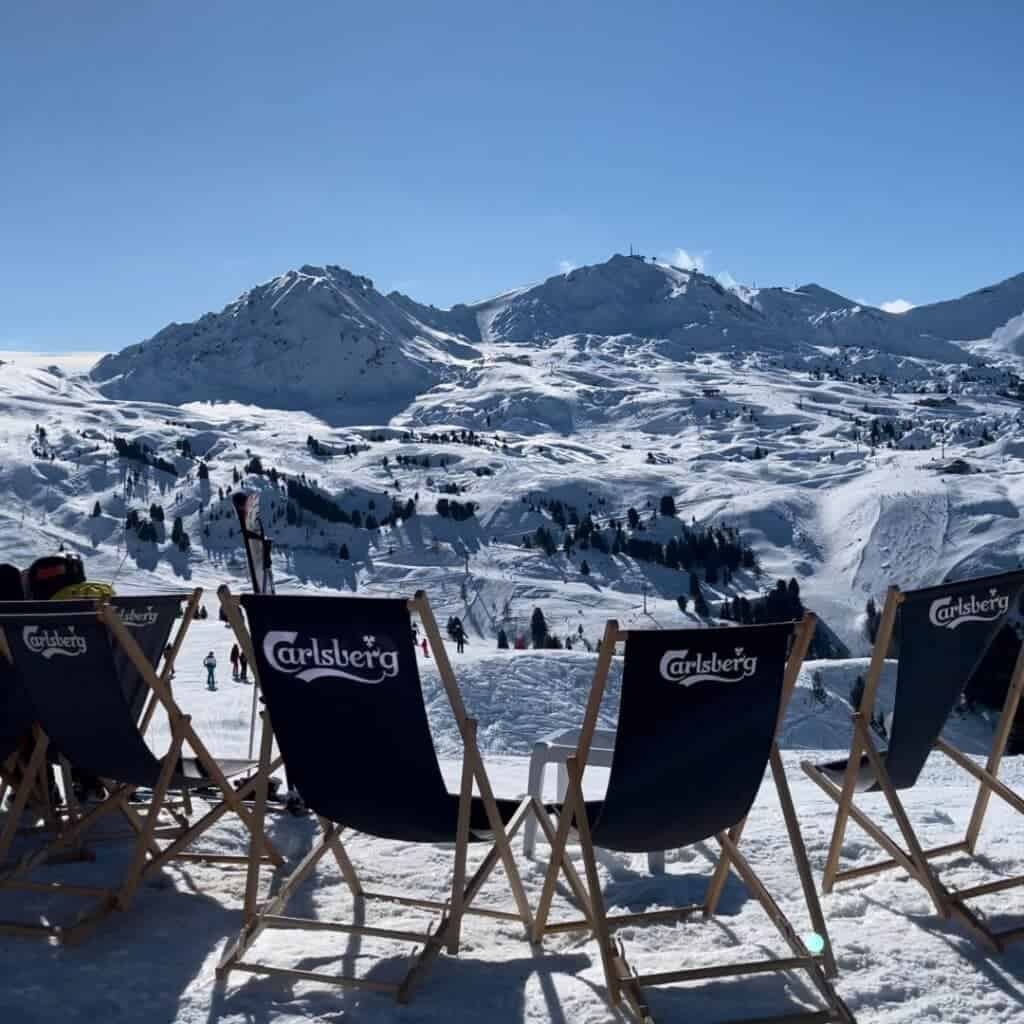 Deck chairs with Carlsberg branding sit on a snowy slope in La Plagne, facing a stunning panorama of sunlit alpine mountains under a clear blue sky. Skiers and snowboarders dot the slopes in the distance, soaking up the crisp February conditions.