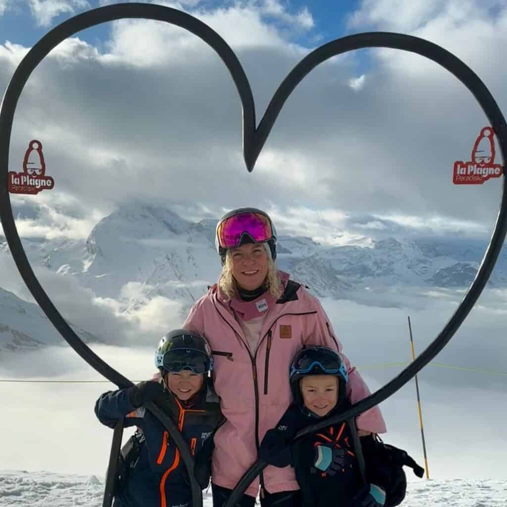 A woman in a pink ski jacket and two children in navy blue ski gear pose in front of a heart-shaped metal frame with "La Plagne Paradiski" signs. Behind them, snow-covered peaks and a sea of clouds stretch across the horizon.