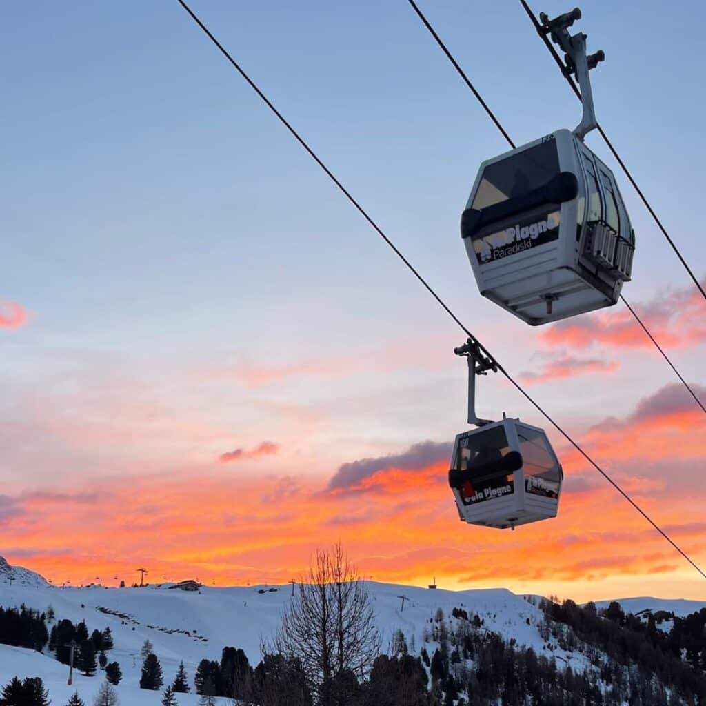 Two La Plagne cable cars soar above snowy slopes during a vivid sunset in the Paradiski area, capturing the serene beauty of La Plagne in March. The sky glows with warm orange and pink tones above a wintery alpine landscape dotted with trees.