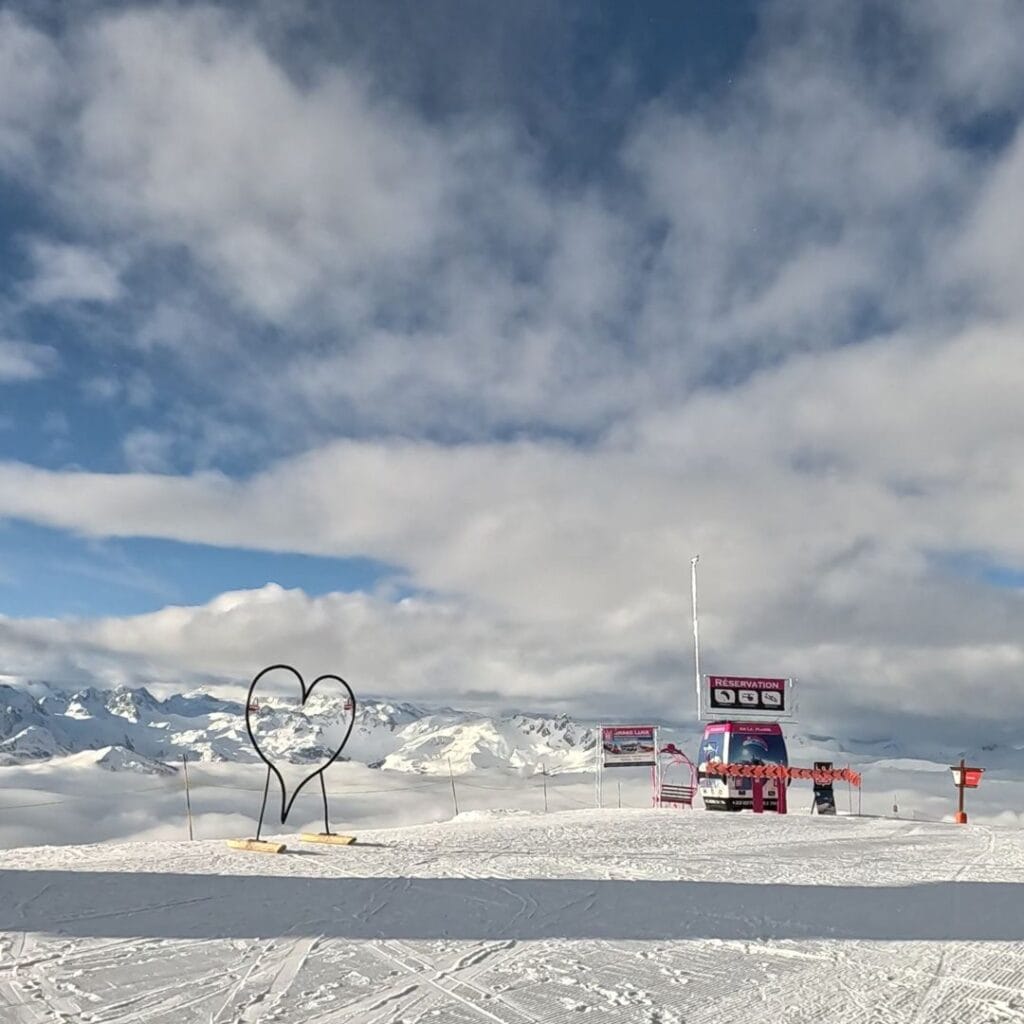 A panoramic view of the La Plagne ski resort with a heart-shaped metal sculpture on the left and a ski station with "Reservation" and other signage in the distance. The untouched snow reflects the bright daylight under a partly cloudy sky.