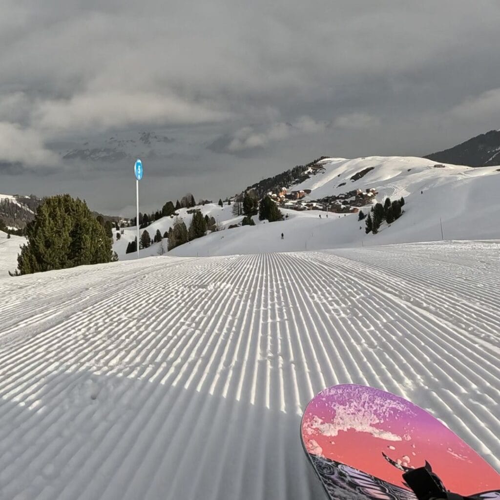 A snowboard with a vibrant pink and purple design rests on the edge of a freshly groomed slope, leading toward a snowy village nestled among rolling hills. A blue slope marker stands to the left, with pine trees dotting the landscape.