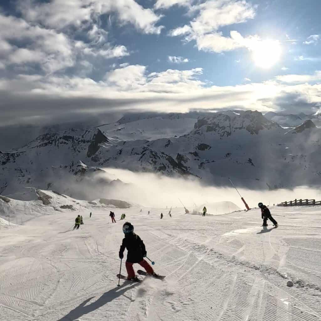 Skiers carve down a sunlit slope in La Plagne, with towering snow-capped peaks and a misty cloud layer in the background. The bright sun shines through the clouds, casting long shadows on the snow.