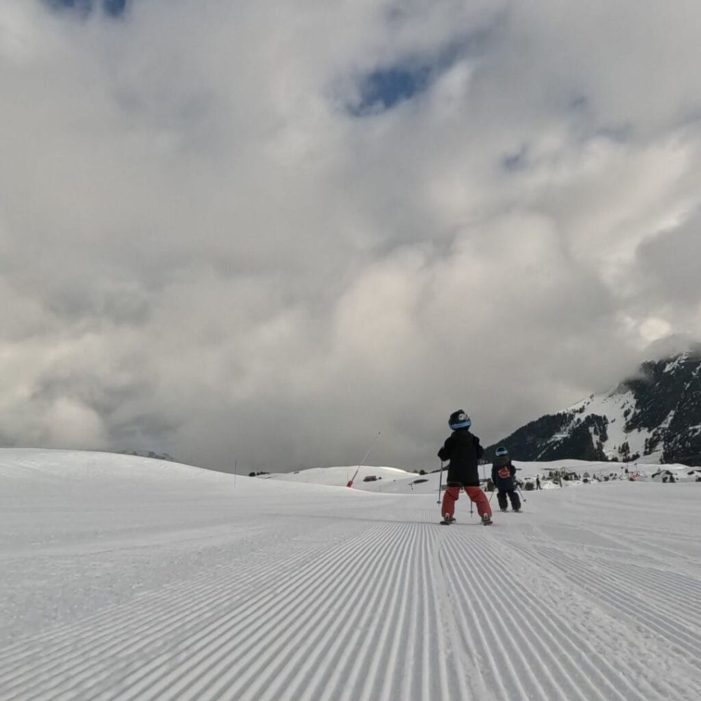 Two young skiers in helmets and ski gear glide down a pristine, groomed slope under a cloudy sky. The vast snowy landscape stretches ahead, with mountain peaks and forested hills in the distance.