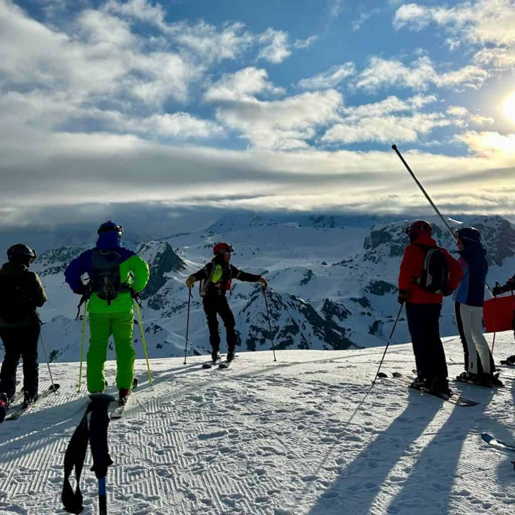 A group of skiers standing on a snow-covered mountain ridge, gazing at the breathtaking view of La Plagne’s peaks under a bright sky with scattered clouds. One skier in a neon green outfit stands prominently, while another gestures toward the slopes.