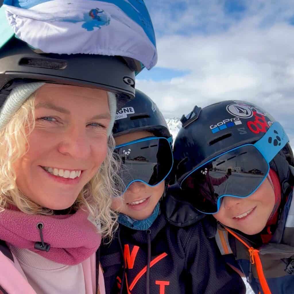 A close-up selfie of a smiling woman with blonde curly hair wearing a ski helmet and a pink scarf, alongside two children in black helmets with ski goggles reflecting the snowy landscape. They are bundled up in winter gear, enjoying their time on the slopes.
