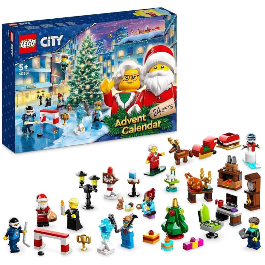 A LEGO City advent calendar set with a snowy holiday scene on the box, including Santa, Mrs. Claus, and minifigures with accessories like gifts, food, and Christmas trees arranged in front of the packaging.
