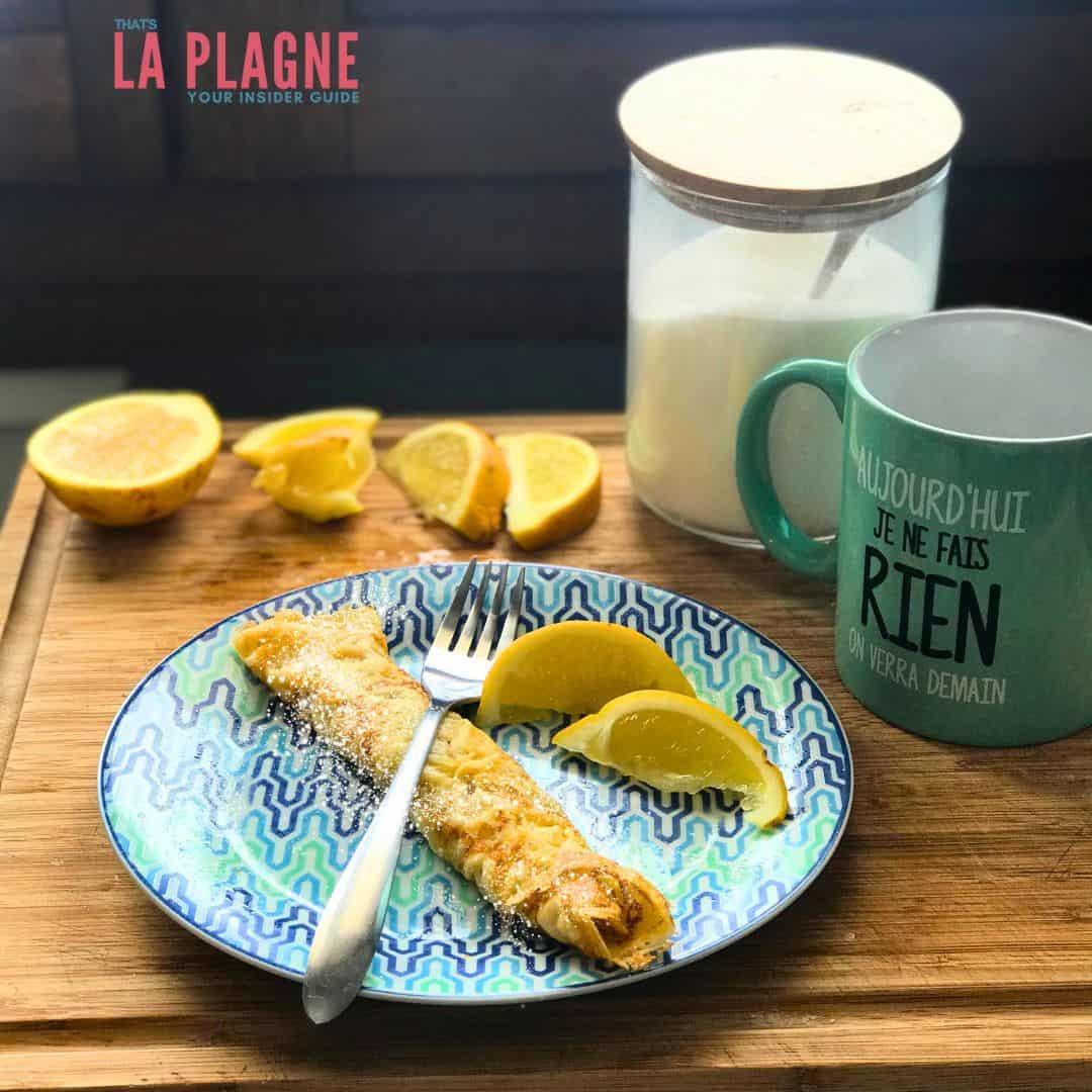 A rolled crepe dusted with powdered sugar is served on a colorful patterned plate with lemon wedges and a fork. The wooden tray also holds a halved lemon, a jar of sugar, and a mint-green mug that reads “Aujourd’hui je ne fais rien, on verra demain” (“Today I’m doing nothing, we’ll see tomorrow”), with a “La Plagne – Your Insider Guide” logo in the corner.