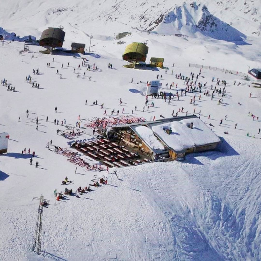 Aerial view of a bustling ski resort scene with skiers and snowboarders gathered near a mountaintop lodge surrounded by snow-covered peaks. The lodge has outdoor seating filled with red lounge chairs, and nearby ski lifts and gondola stations serve the busy slope area.