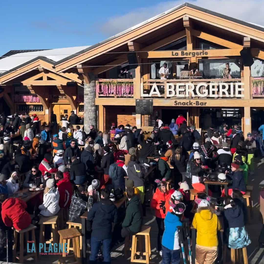 A lively crowd gathers outside La Bergerie Snack-Bar, a wooden alpine-style restaurant with a snow-covered roof. People in colorful winter clothing are standing, sitting on stools, and socializing under a clear blue sky. The scene suggests a popular après-ski spot in a mountain resort.
