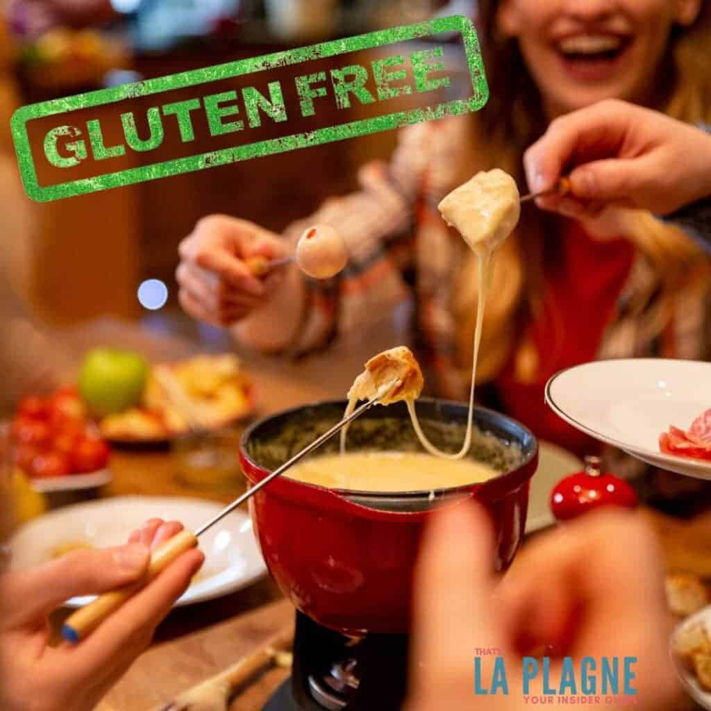 A lively gathering of friends enjoying a gluten-free cheese fondue in a red pot. Hands hold skewers with bread, cheese, and other dippables, stretching gooey melted cheese. A bold green 'GLUTEN FREE' stamp overlays the image, with 'La Plagne' branding in the corner, suggesting a cozy, food-friendly experience in the region.