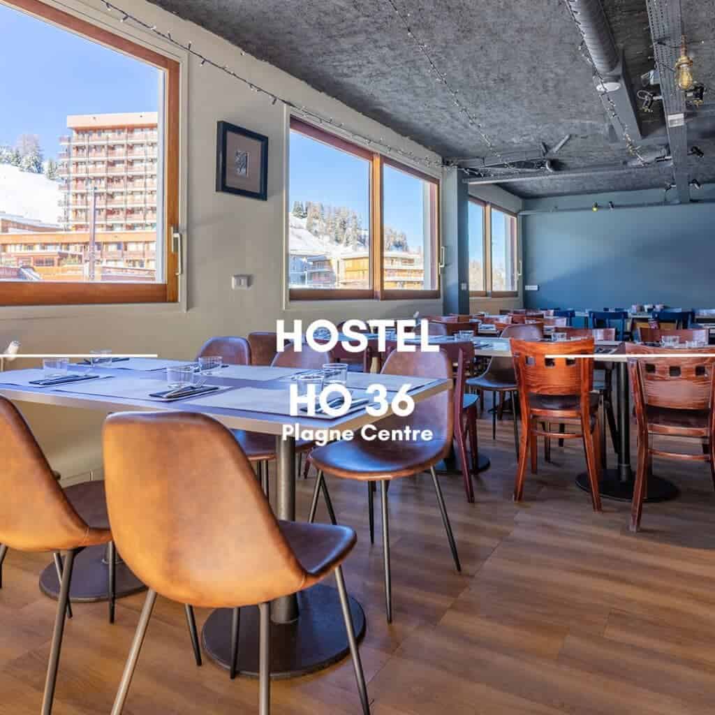 A sunlit dining room at Hostel HO36 in Plagne Centre, with modern wooden tables and chairs, and large windows offering views of snowy buildings and trees outside.