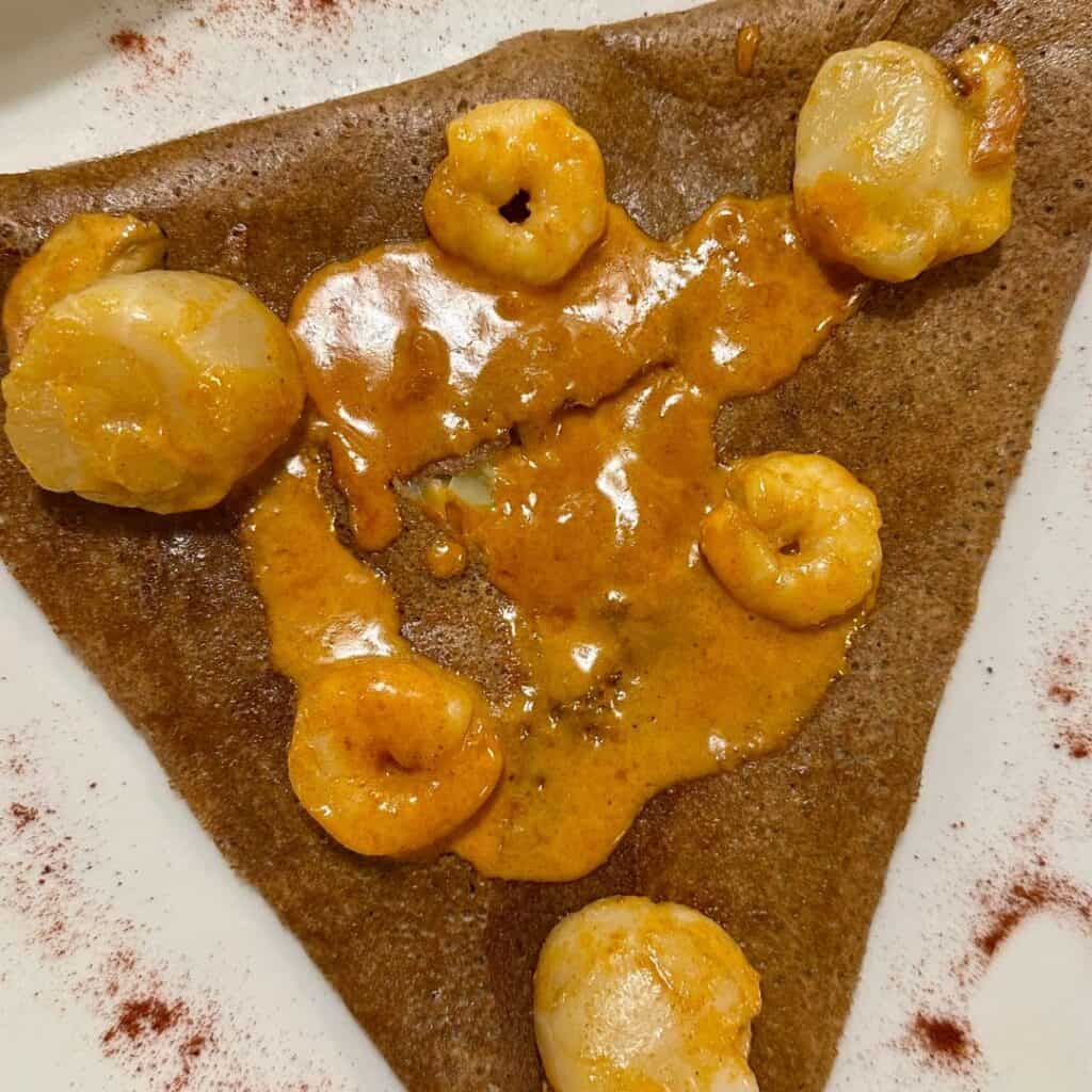 Close-up of a savory buckwheat galette topped with shrimp and scallops in a creamy orange sauce, folded into a triangle on a white plate with paprika garnish.