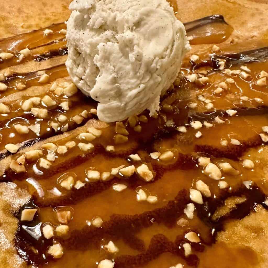 Close-up of a sweet crepe drizzled with caramel and chocolate sauces, sprinkled with chopped nuts, and topped with a generous scoop of vanilla ice cream.