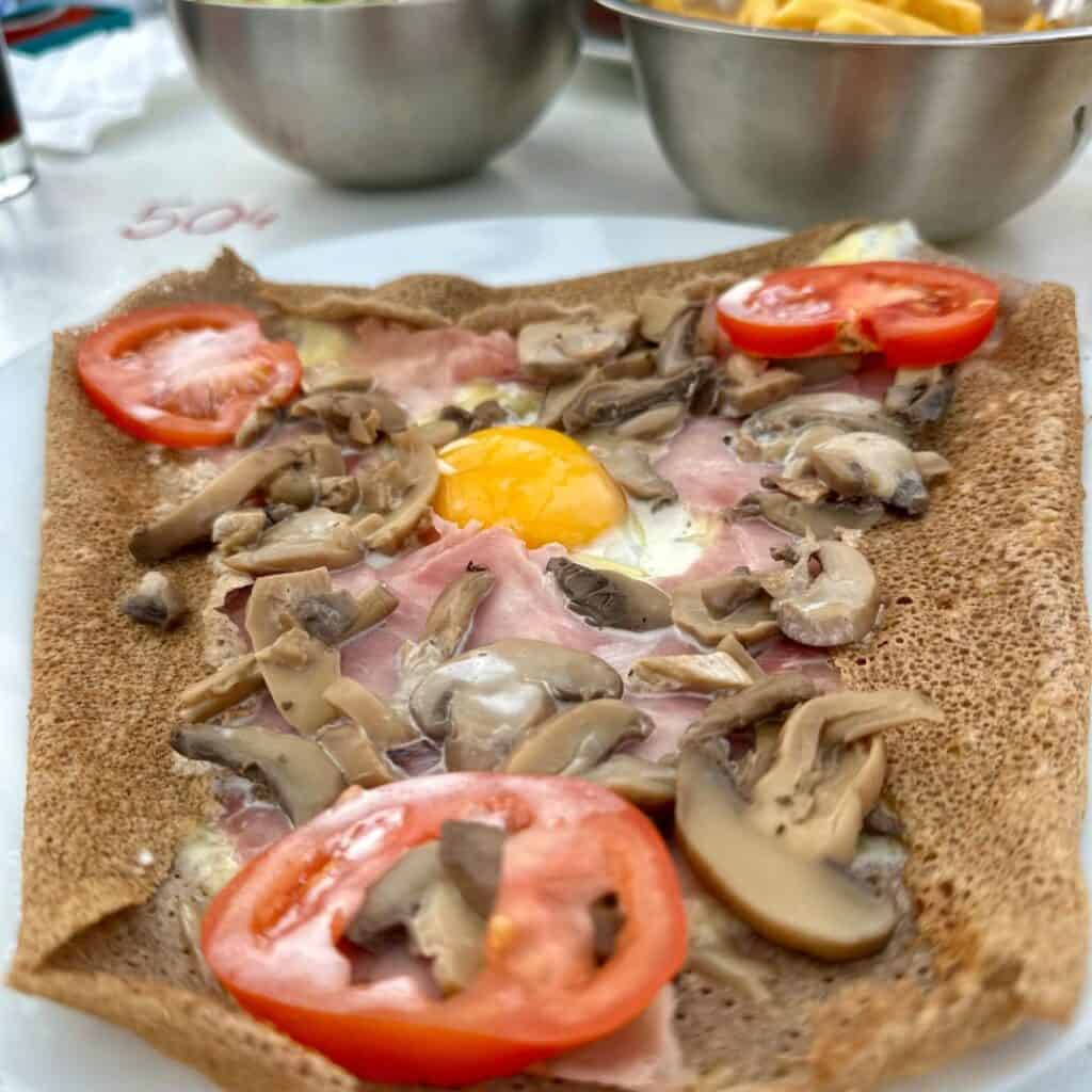 Close-up of a savory buckwheat galette topped with sliced tomatoes, mushrooms, ham, and a sunny egg yolk in the center, served on a white plate with bowls of fries and salad in the background.