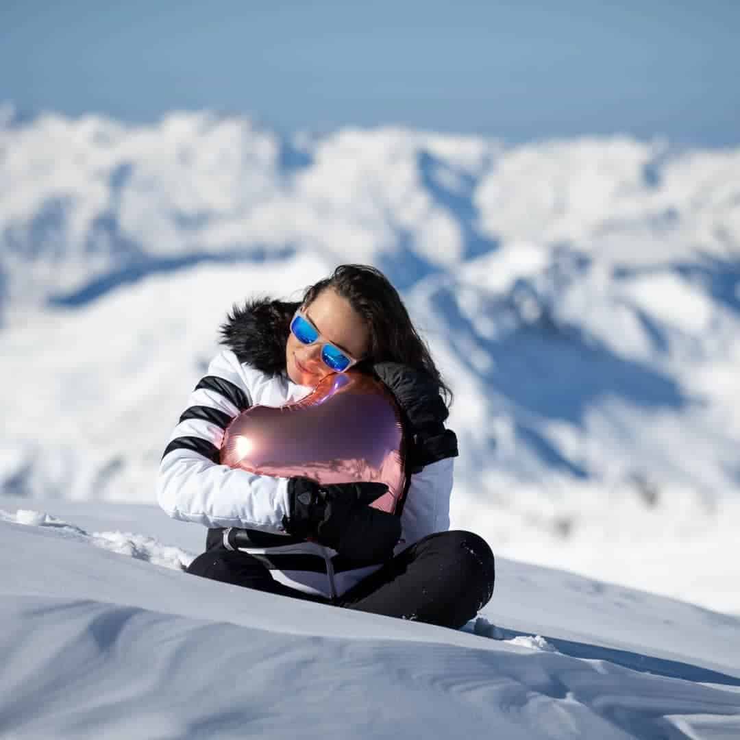 A woman in a white and black winter jacket sits in the snow hugging a shiny, rose gold heart-shaped balloon. She’s wearing reflective blue sunglasses, and behind her is a breathtaking view of snow-covered mountains under a clear blue sky.