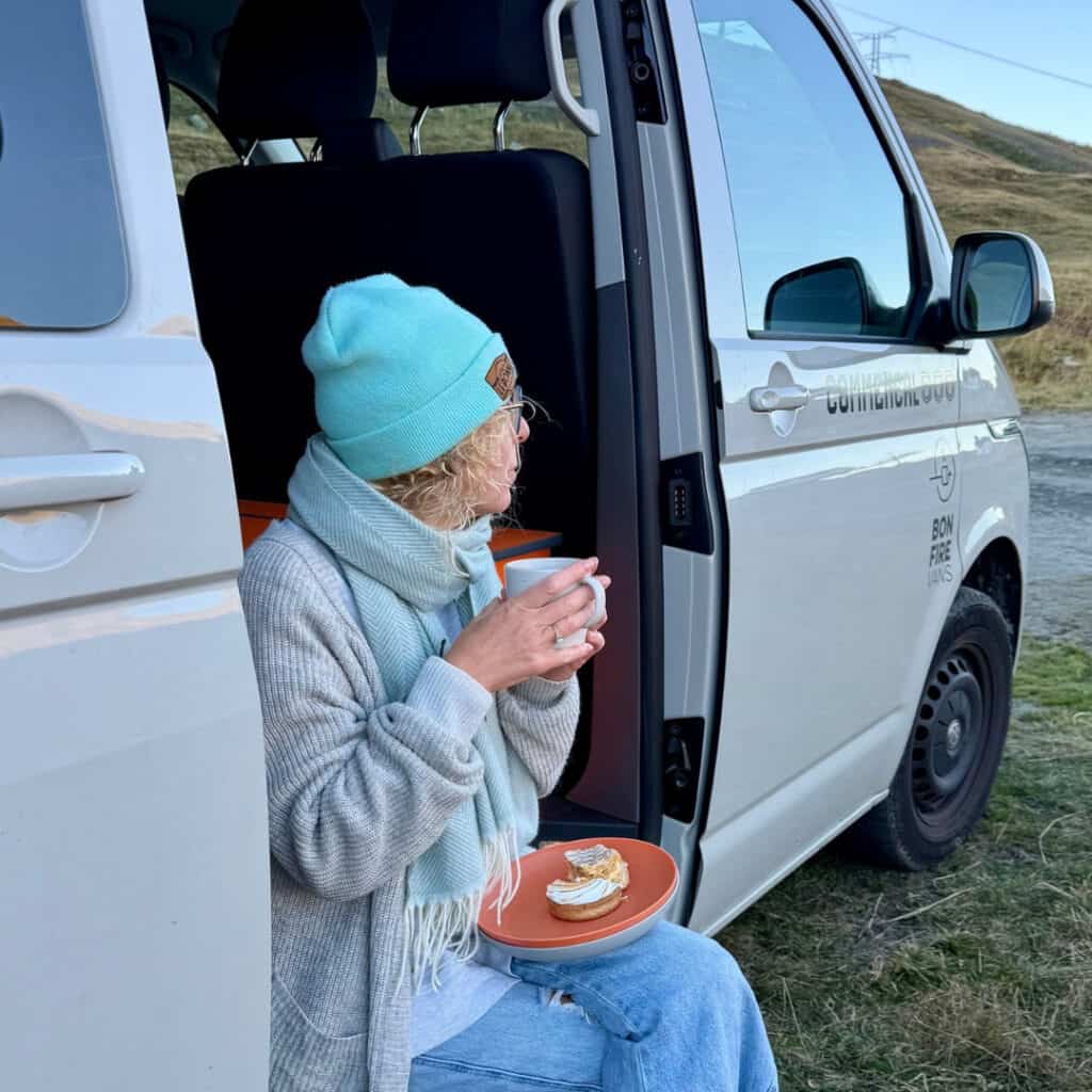 Woman sitting in the open doorway of a white campervan in La Plagne, holding a hot drink with a pastry on her lap in an Alpine mountain setting.