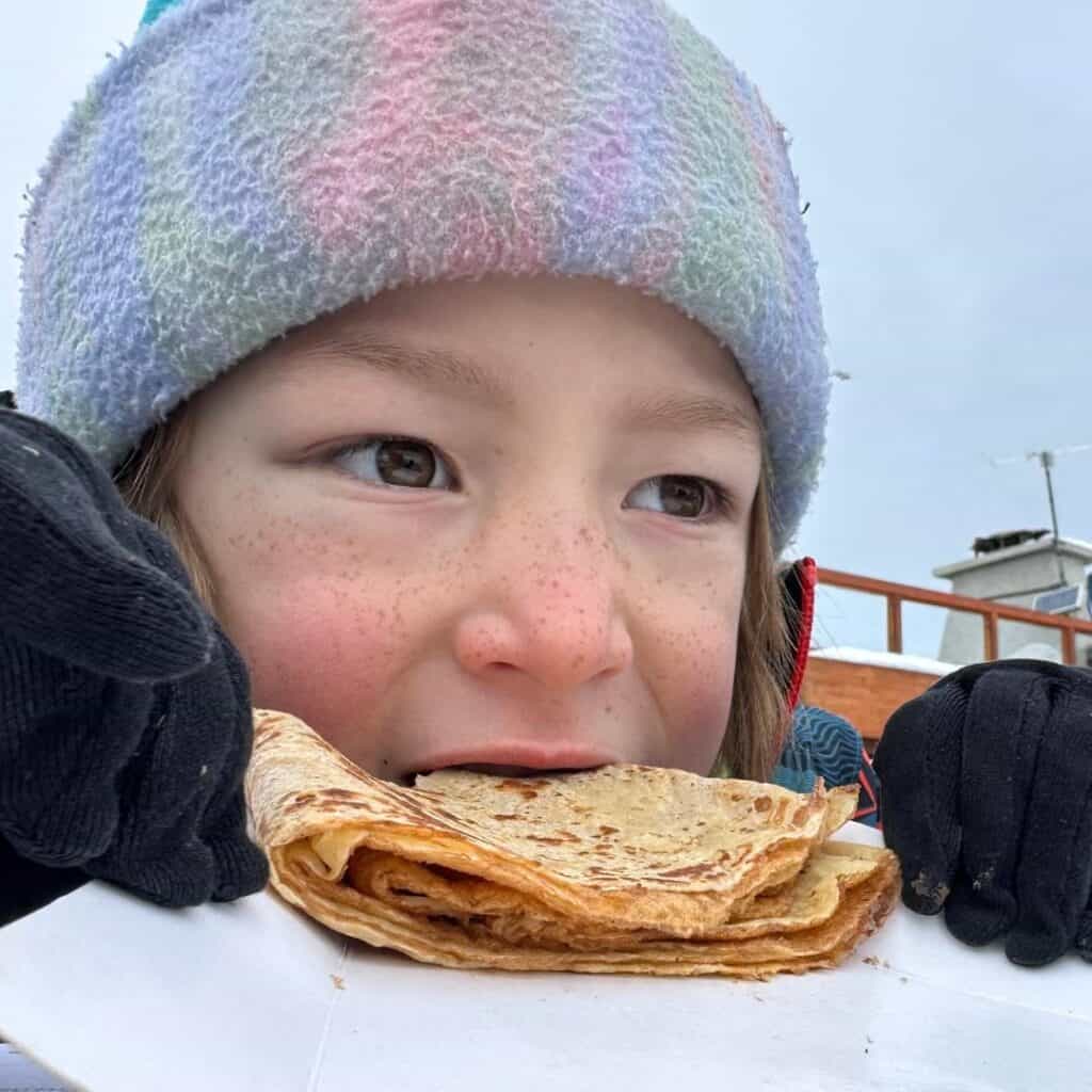 Close-up of a child in a pastel-striped winter hat and black gloves taking a bite of a folded crepe, with a snowy outdoor setting in the background. The child’s cheeks are rosy, and they look content and cozy.