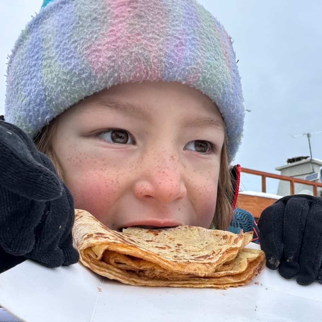 Close-up of a child in a pastel-striped winter hat and black gloves taking a bite of a folded crepe, with a snowy outdoor setting in the background. The child’s cheeks are rosy, and they look content and cozy.