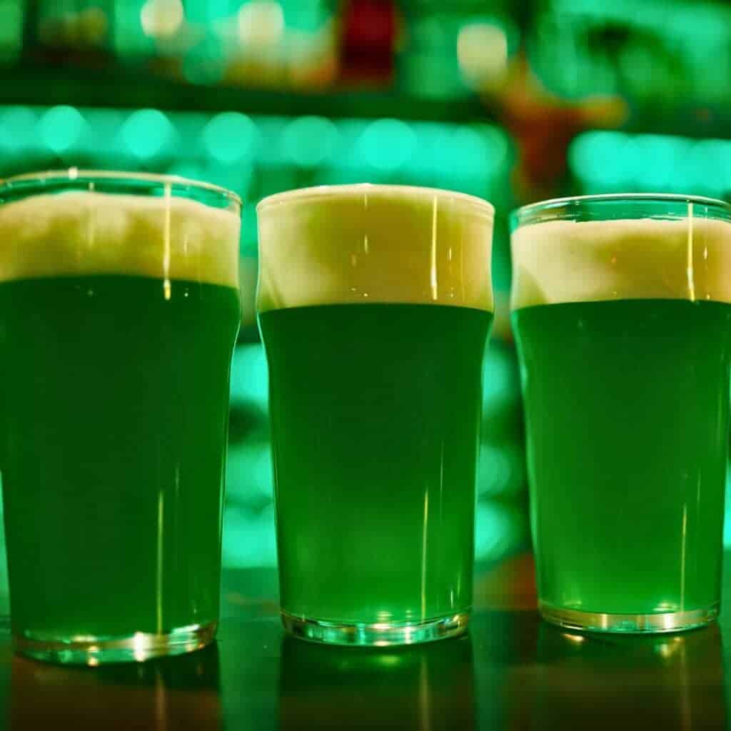 Three pint glasses filled with vibrant green beer sit on a bar counter, topped with a frothy white head. The background glows with blurred green lights, creating a festive atmosphere. Perfect for celebrating St. Patrick's Day.