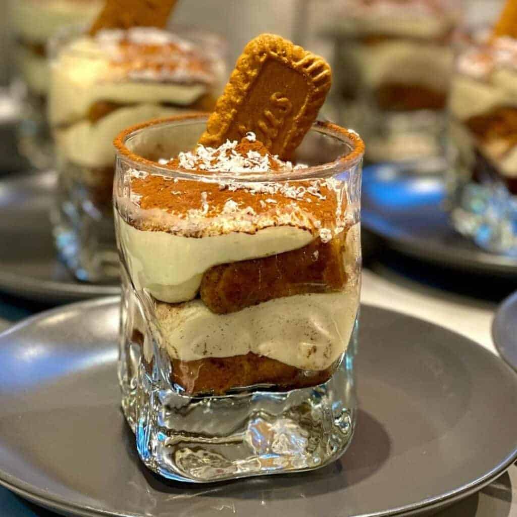 Alpoholics catered ski chalets La Plagne. A glass cup filled with layers of creamy mascarpone and biscuit, dusted with cocoa powder and topped with a Biscoff cookie. The dessert is served on a grey plate with similar cups in the background.