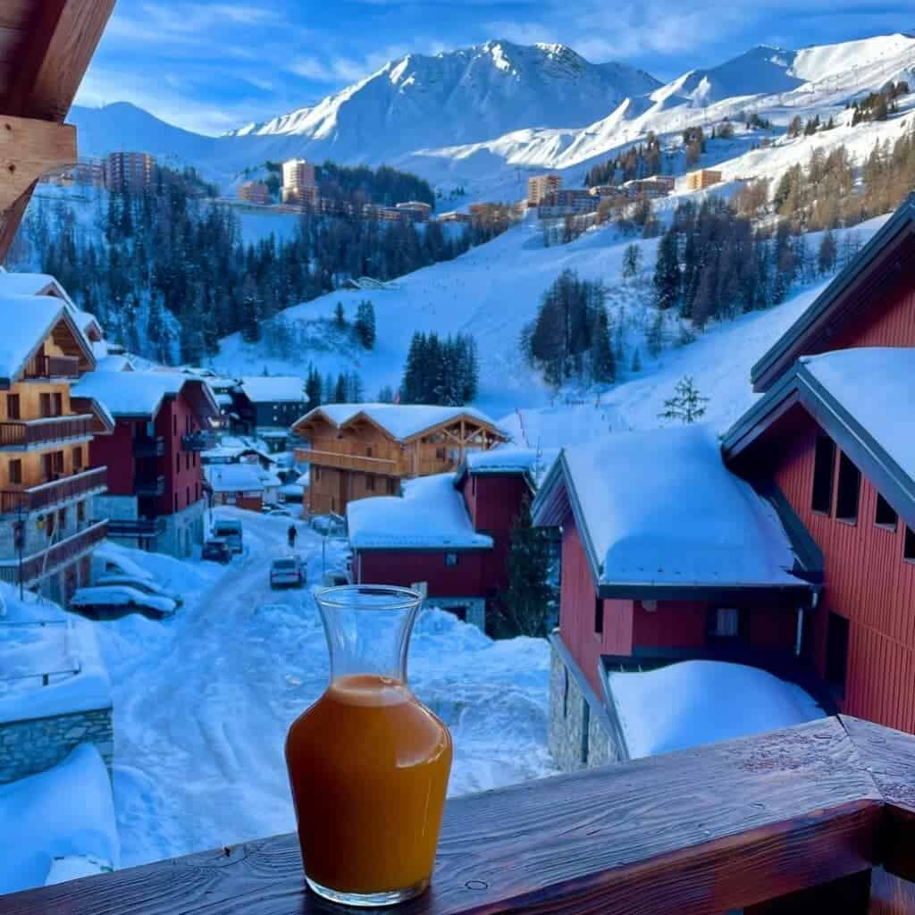 Alpoholics catered ski chalets La Plagne. A glass carafe filled with orange juice sits on a wooden balcony railing overlooking a snowy alpine village. The background features snow-covered wooden chalets and distant mountain peaks under a bright blue sky.