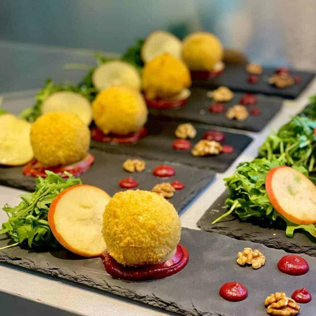 Alpoholics catered ski chalets La Plagne. A gourmet appetizer featuring golden, breaded cheese balls served on black slate plates. Each portion is accompanied by fresh arugula, thin apple slices, walnuts, and small dollops of red sauce, creating a visually elegant presentation.