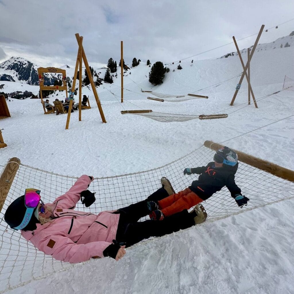 A woman and child relax on a snow-side hammock at the Buffalo Fun Slope picnic area in La Plagne, surrounded by snowy mountains, pine trees, and wooden loungers—one of the most playful and family-friendly picnic spots in the resort.