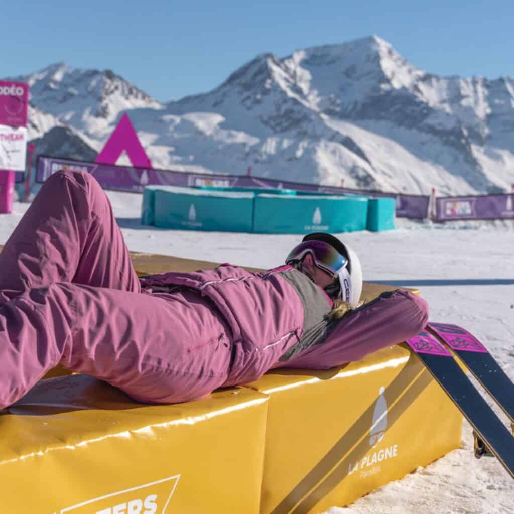 A skier in a pink snowsuit lounges on a bright yellow platform at Riders Nation snow park in La Plagne, enjoying a laid-back picnic break with stunning alpine scenery all around.