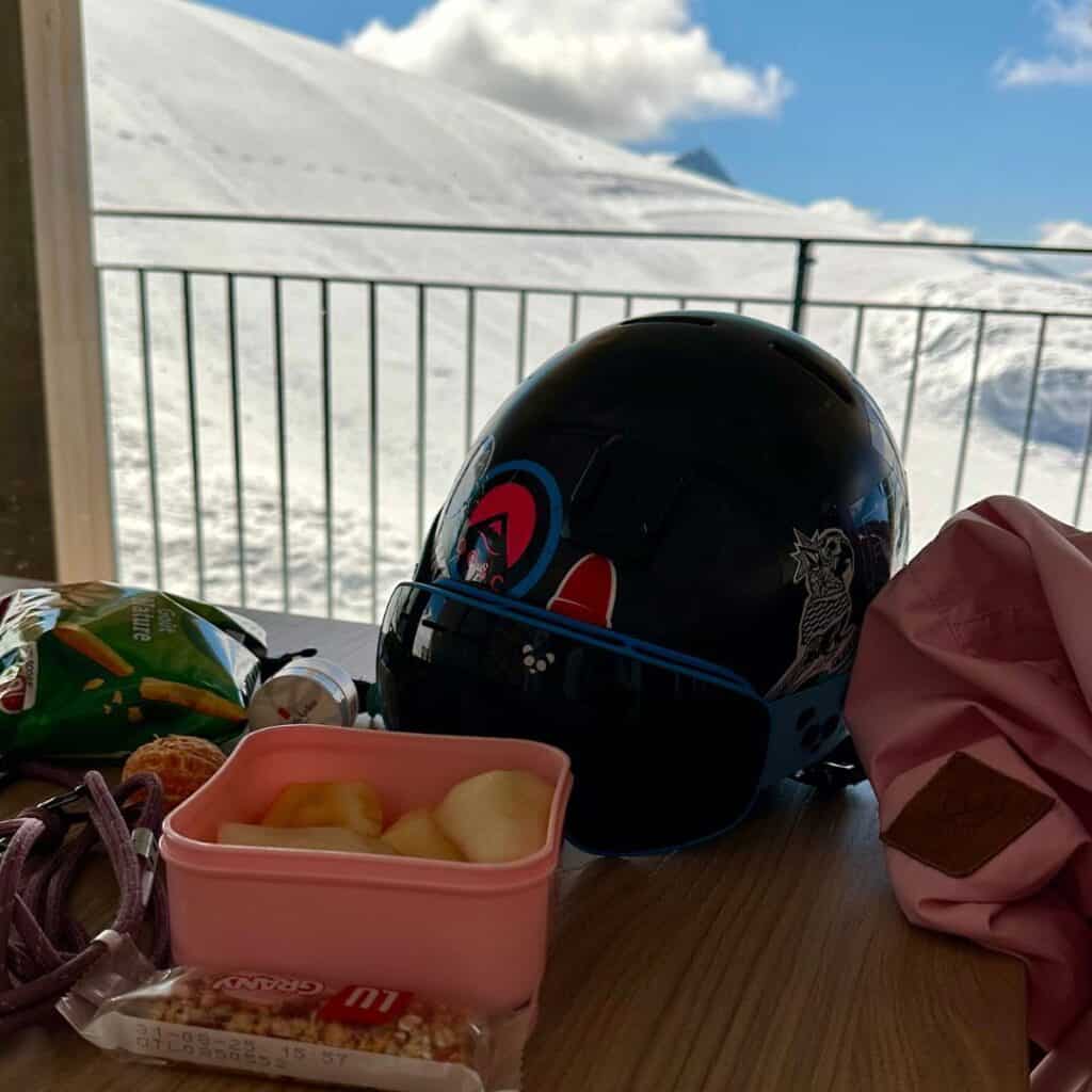 Ski helmet, jacket, snacks, and fruit laid out on a table at Roche de Mio, one of the most convenient La Plagne picnic spots with indoor warmth and snowy mountain views just outside.